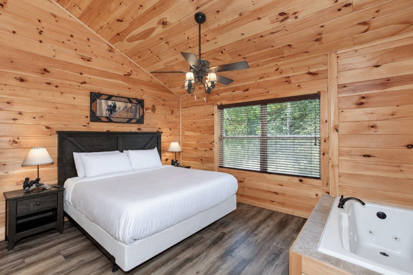 Une chambre à coucher dans une cabane en bois, comprenant un lit, des tables de chevet, un ventilateur de plafond, une fenêtre avec des stores et une baignoire.