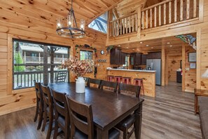 A wooden cabin interior with a dining area, kitchen, and a balcony. - Creekside Lodge | Hot Tub, Grill, Fireplace, GAMES (Pigeon Forge)