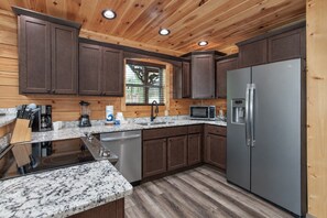 A modern kitchen with wooden cabinets, granite countertops, stainless steel appliances, and a window providing natural light. - Creekside Lodge | Hot Tub, Grill, Fireplace, GAMES (Pigeon Forge)