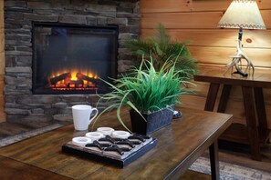 A cozy room with a stone fireplace, a wooden table with a tray of cookies, a white mug, and a potted plant. - Creekside Lodge | Hot Tub, Grill, Fireplace, GAMES (Pigeon Forge)