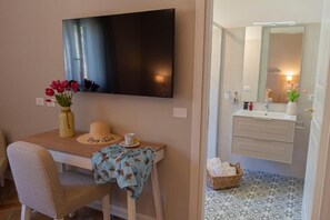 A room with a television mounted on the wall, a desk with a vase of flowers, a hat, and a cup. There is a bathroom with a mirror, sink, and patterned floor tiles. - Villa Cammà with private pool (Mirto)