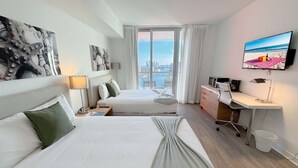A hotel room with two beds, a desk, a chair, a TV, and a view of the city and water. - Comfortable BW Studio - Pool & Bay View (Hallandale Beach)