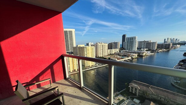A balcony with a red wall, a glass railing, and a view of a city skyline across a body of water. - Comfortable BW Studio - Pool & Bay View (Hallandale Beach)