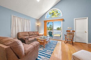 A living room with a brown leather sofa, a glass coffee table, a rocking chair, and a view of a balcony with a railing. - Colington Harbour 34: Soundfront with Private Dock (Colington)
