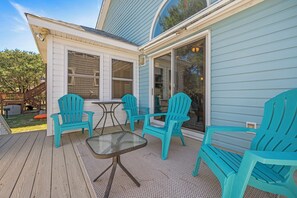 A deck with a glass-top table and four turquoise chairs, a wooden deck, and a house with light blue siding. - Colington Harbour 34: Soundfront with Private Dock (Colington)