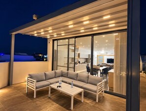 A modern outdoor seating area with a sofa and table, a glass door leading to a kitchen, and a view of the cityscape. - Rokna corner penthouse - 240sqm with sea view in Xaghra Gozo (Ix-Xagħra)
