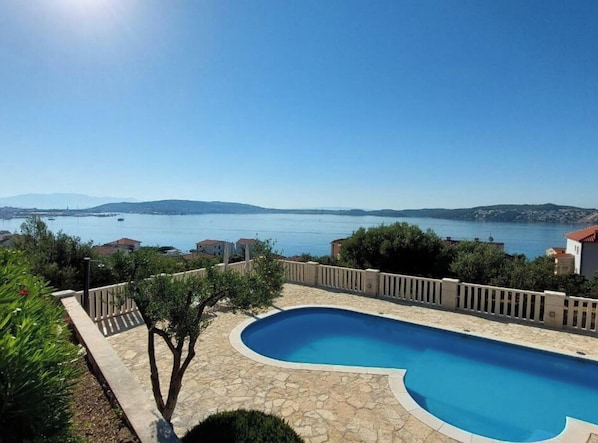 A swimming pool with a curved edge, surrounded by a stone deck and a wooden fence. - Villa in Seget Donji With Swimming Pool (Seget)