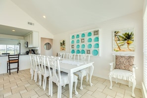 Un área de comedor con una mesa y sillas blancas, una cocina al fondo y una pared con un cuadro de una playa tropical y platos color turquesa.