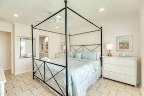 A four-poster canopy bed with a white and blue floral bedspread, a white dresser with drawers, a framed picture on the wall, and a ceiling fan.