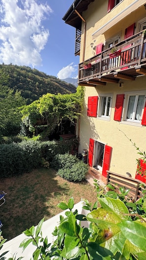 A multi-story building with red shutters and a balcony, surrounded by lush greenery and a mountainous backdrop.