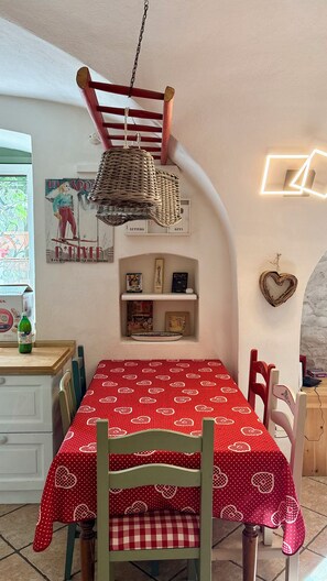 A dining area with a table set for two, a hanging light fixture, and a wall-mounted shelf with books and decorative items.