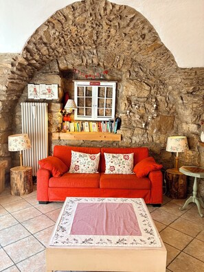 A cozy room with a stone wall, a red sofa, a wooden table, and a 'Welcome' sign above a window.