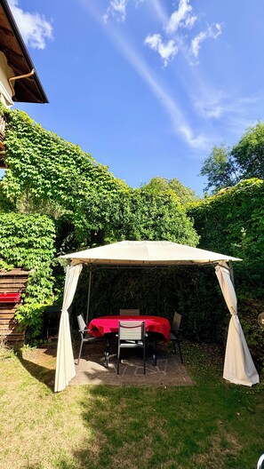 A covered outdoor dining area with a table and chairs, surrounded by lush greenery and a clear blue sky. - Sopramonte Apartment and Annex (Sopramonte)