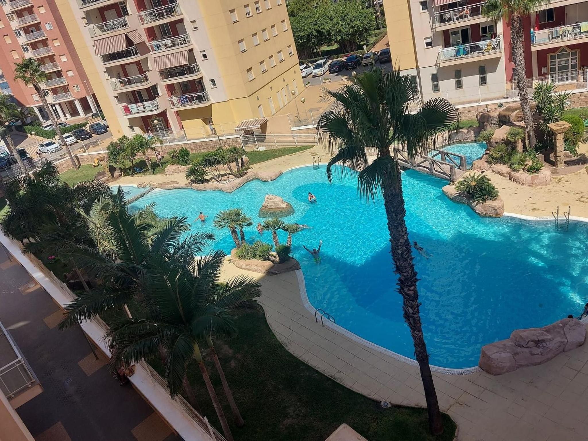 Una piscina rodeada de palmeras y un puente de piedra, con vista a un edificio de varios pisos al fondo.