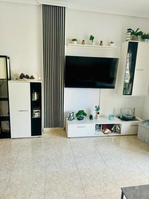 Una sala de estar moderna con un televisor de pantalla plana montado en la pared, un mueble blanco para equipos electrónicos y un armario pequeño con espejo.