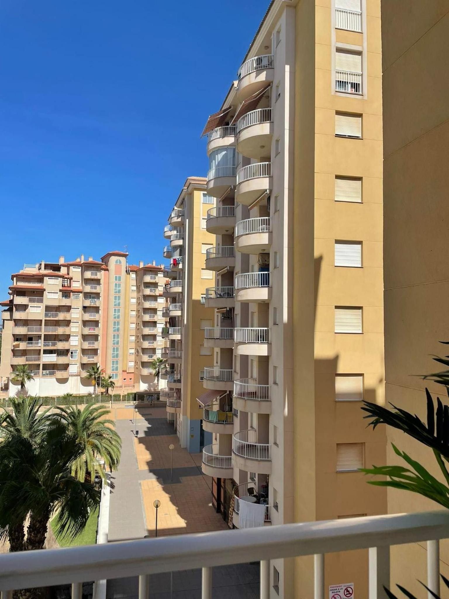 Un edificio residencial de varios pisos con balcones, cielo despejado y palmeras en primer plano.