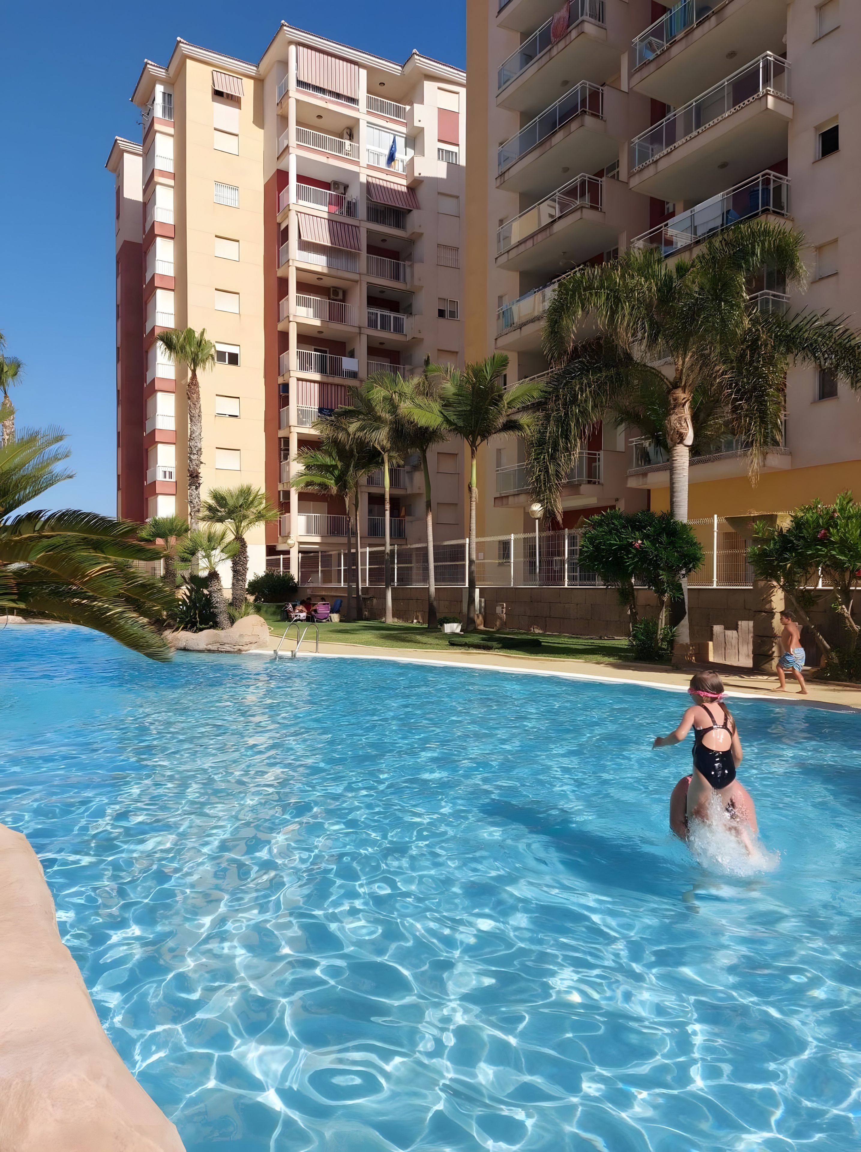 Ein Pool mit klarem Wasser, umgeben von Palmen und einem mehrstöckigen Gebäude mit Balkonen.