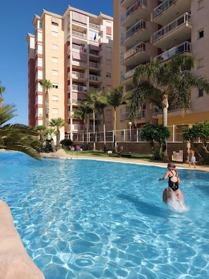 Una piscina con agua cristalina, rodeada de palmeras y un edificio de varios pisos con balcones.