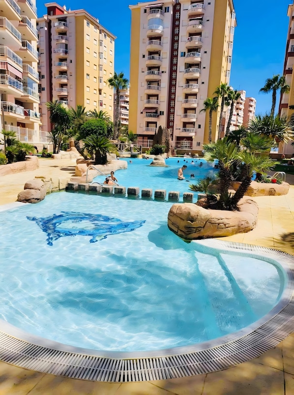 Piscina com formações rochosas e palmeiras, cercada por edifícios altos.