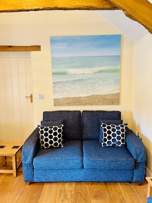 Een kamer met een blauwe bank, twee gestreepte kussens, een houten tafel en een schilderij met een strandthema aan de muur.