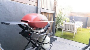 A Weber Q grill with a gas tank, positioned on a concrete patio with a wooden fence and a white chair in the background.