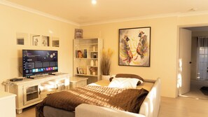 A modern bedroom with a TV on a wooden stand, a bed with a brown quilt, a bookshelf, and a painting on the wall.