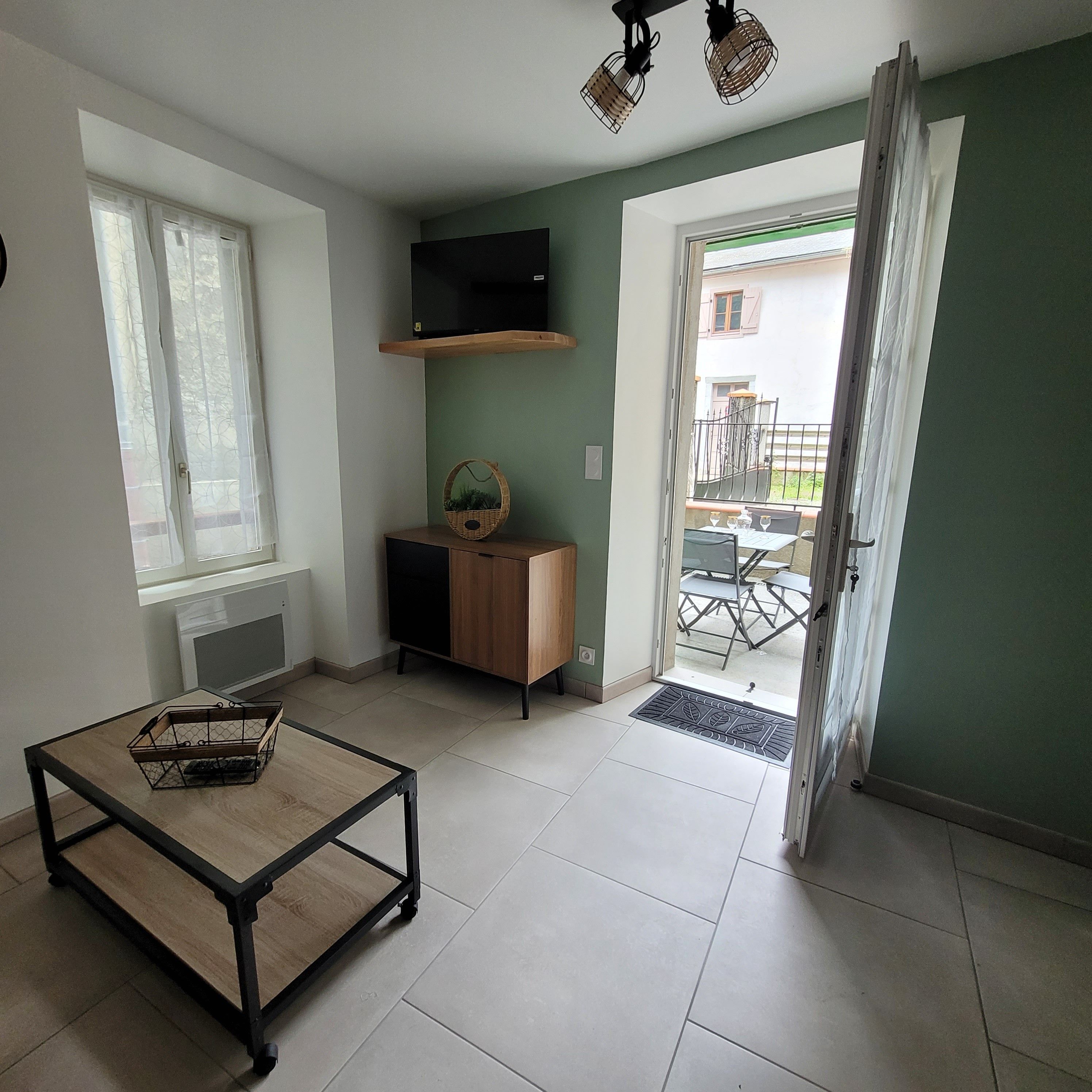 A room with a TV on a shelf, a small cabinet, a table with a basket, and a view of the outside balcony.