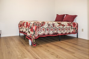 A bed with a patterned bedspread and two pillows, a wooden floor, and white walls.