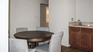 Uma sala de jantar com uma mesa redonda e quatro cadeiras, uma cozinha com pia e armários de madeira, e uma porta de entrada para outro cômodo.