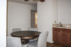 Un comedor con una mesa redonda y cuatro sillas, una cocina con un lavaplatos y armarios de madera, y una puerta que da a otra habitación.