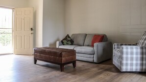 Uma sala de estar com um sofá cinzento, uma poltrona xadrez e um banquinho marrom.