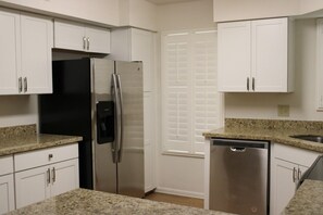 A kitchen with white cabinets, a stainless steel refrigerator, a dishwasher, and a window with blinds.
