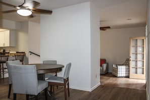 A dining area with a round table, four chairs, a ceiling fan, and a kitchen in the background.