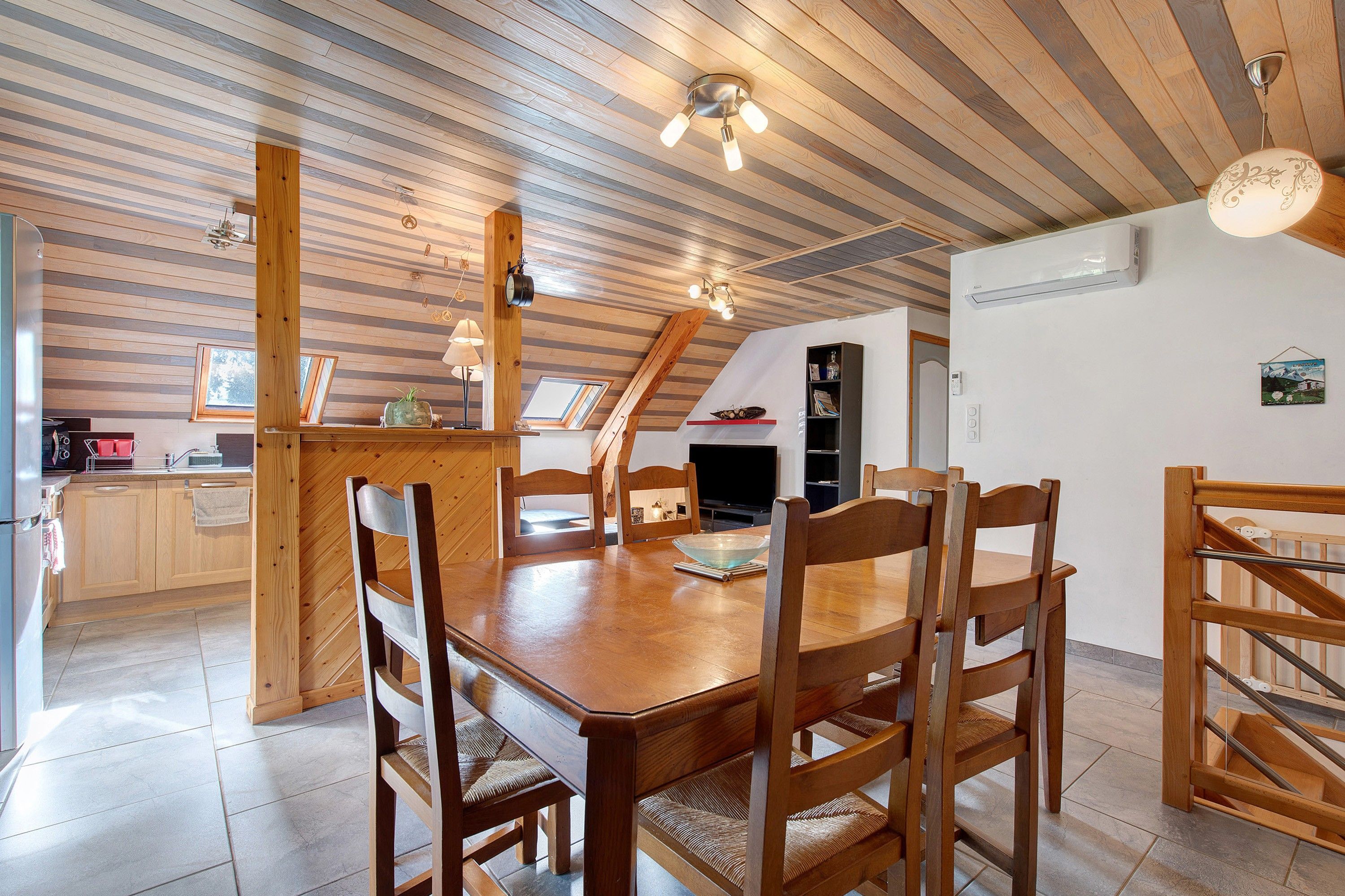 Et spiseområde med et træbord og stole, et køkken med et køleskab og skabe, samt et skråt loft med træbjælker.