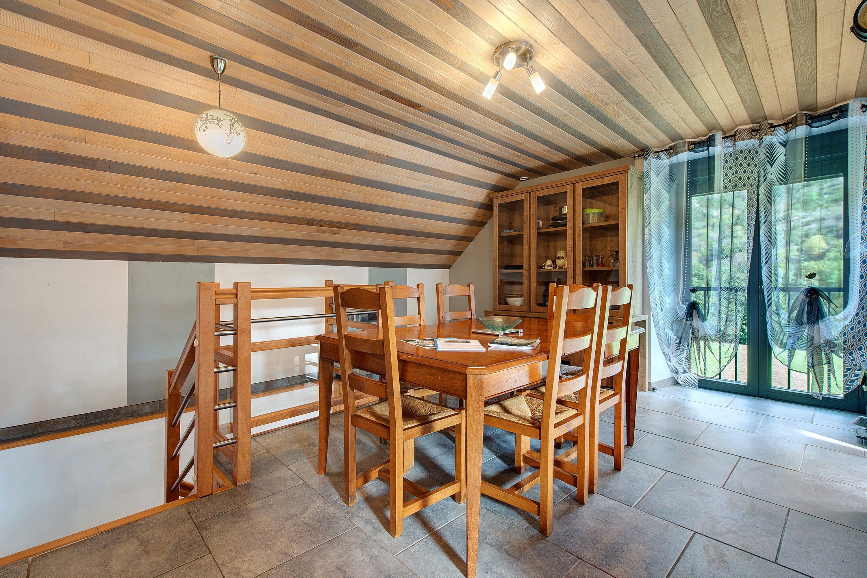 A dining area with a wooden table and chairs, a staircase, and a view of the outdoors through a window.
