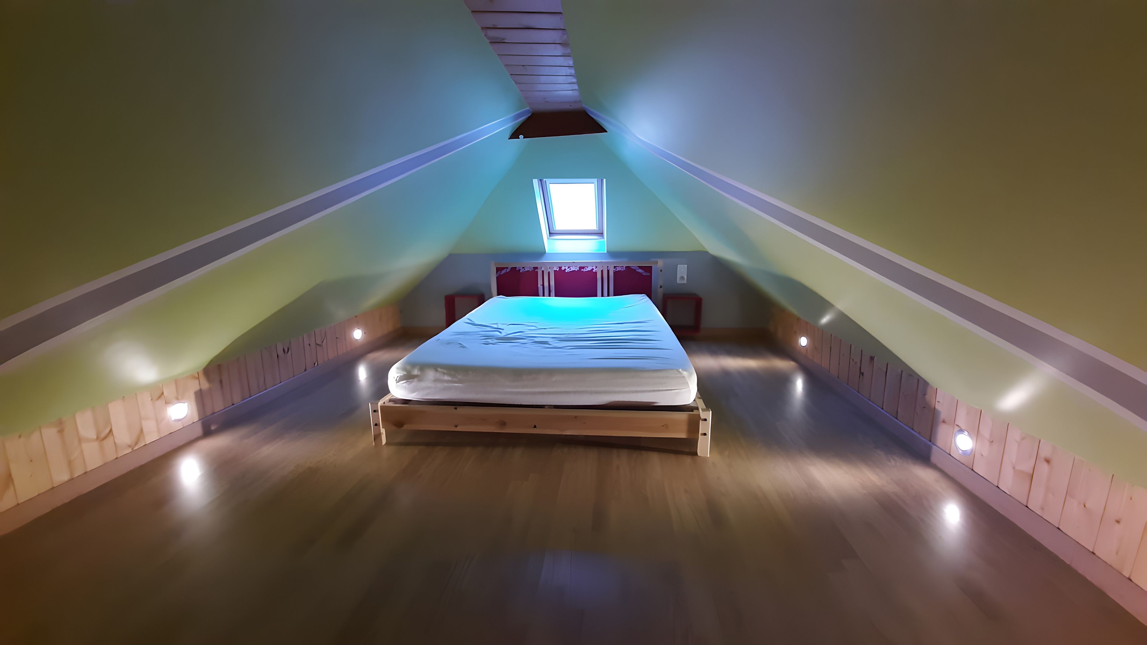 Una habitación con techo inclinado, una cama, un ventanal y un tragaluz pequeño.