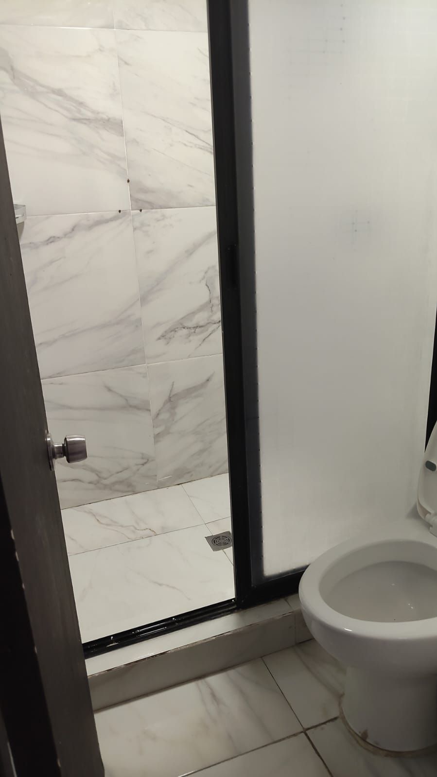 Un baño con zona de ducha con azulejos de mármol blanco, un inodoro y una puerta con pomo metálico.