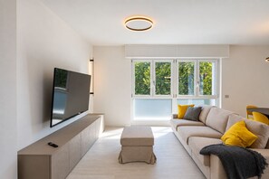 A modern living room with a flat-screen TV, a sofa with yellow cushions, and a large window with a view of greenery. - Lake Iseo - Waterfront House | Sarnico (Sarnico)