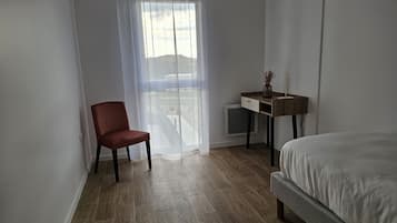 A bedroom with a bed, a chair, a desk with a vase, and a window with sheer curtains.