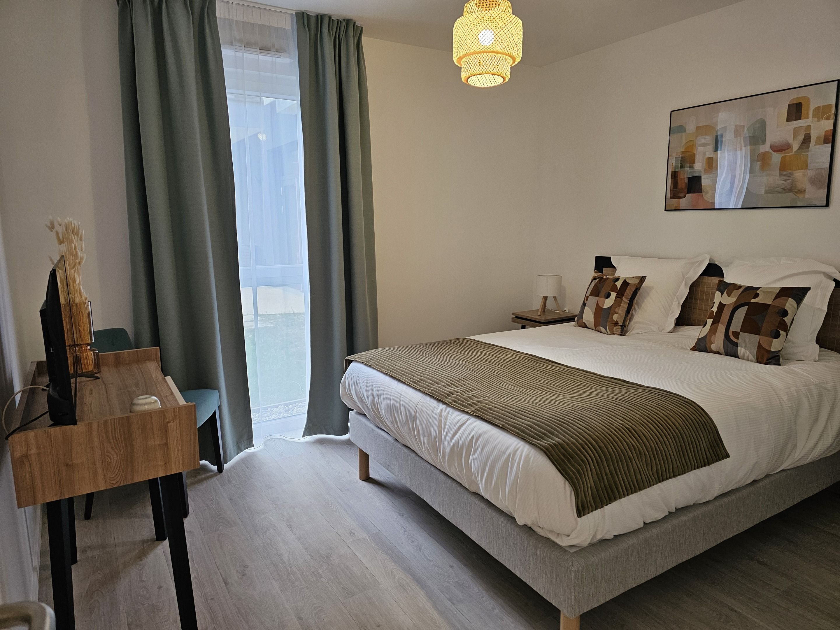 Un dormitorio con cama, mesita de noche, lámpara, un cuadro y vistas a través de la ventana.