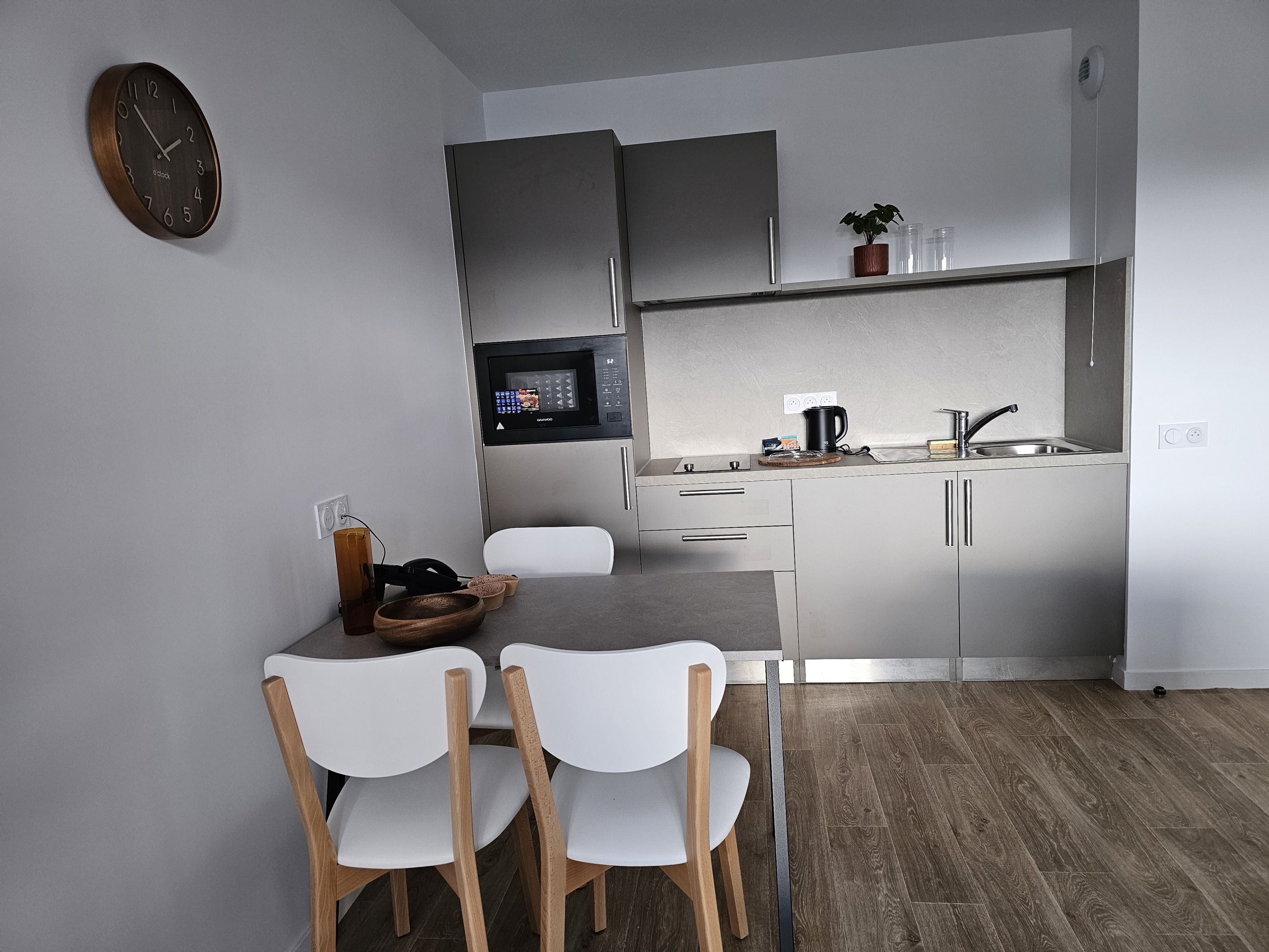 Een moderne keuken met een magnetron, spoelbak en kastjes. Er is een eettafel met stoelen en een klok aan de muur.
