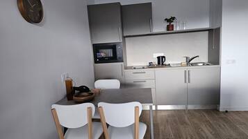Een moderne keuken met een magnetron, spoelbak en kastjes. Er is een eettafel met stoelen en een klok aan de muur.