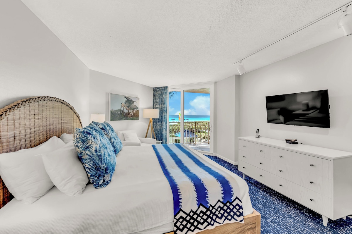A modern hotel room with a bed, a TV, a dresser, and a view of the ocean.