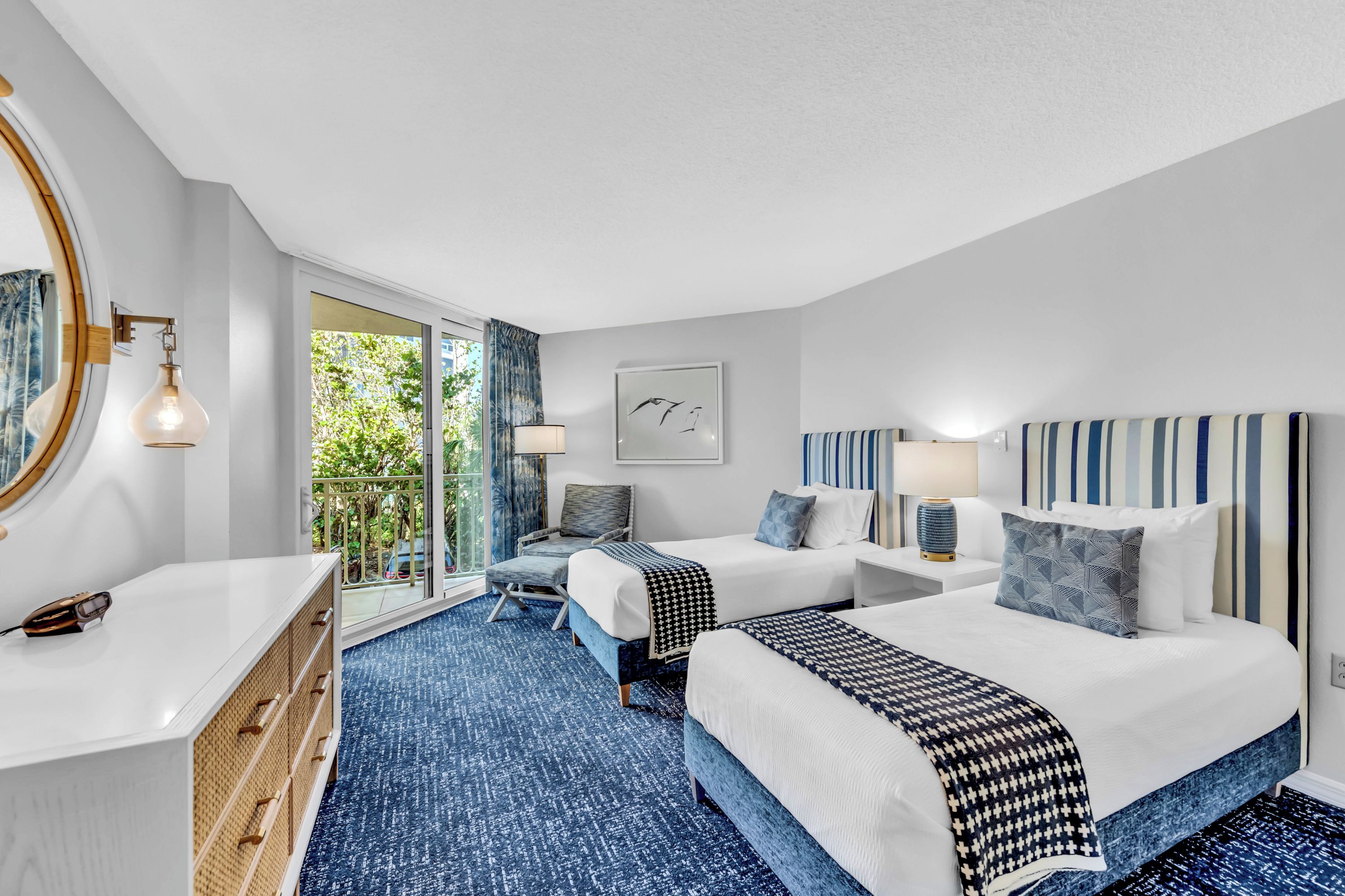 A hotel room with two beds, a large window, a balcony, and a view of greenery.