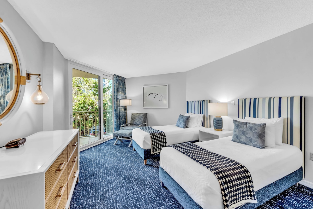A hotel room with two beds, a large window, a balcony, and a view of greenery.