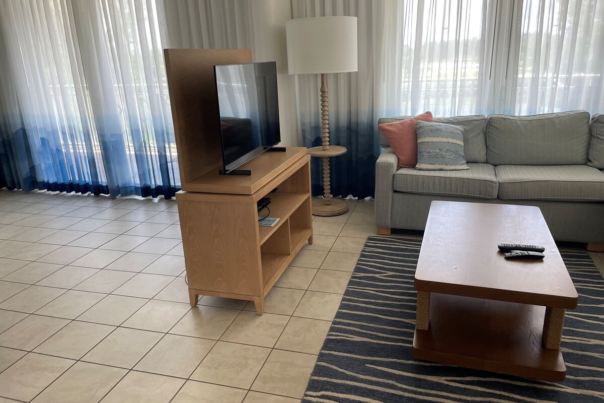 A living room with a television on a wooden stand, a sofa with cushions, a coffee table, and a lamp.