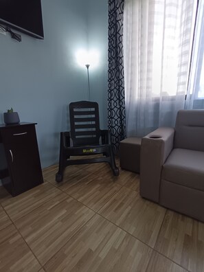 Ein Wohnzimmer mit einer Couch, einem kleinen Tisch, einem Sessel, einem Schrank und einem an der Wand befestigten Fernseher.