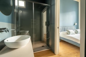 Une salle de bain moderne avec une douche en verre, un lavabo blanc et un lit avec du linge de lit blanc dans la pièce adjacente.