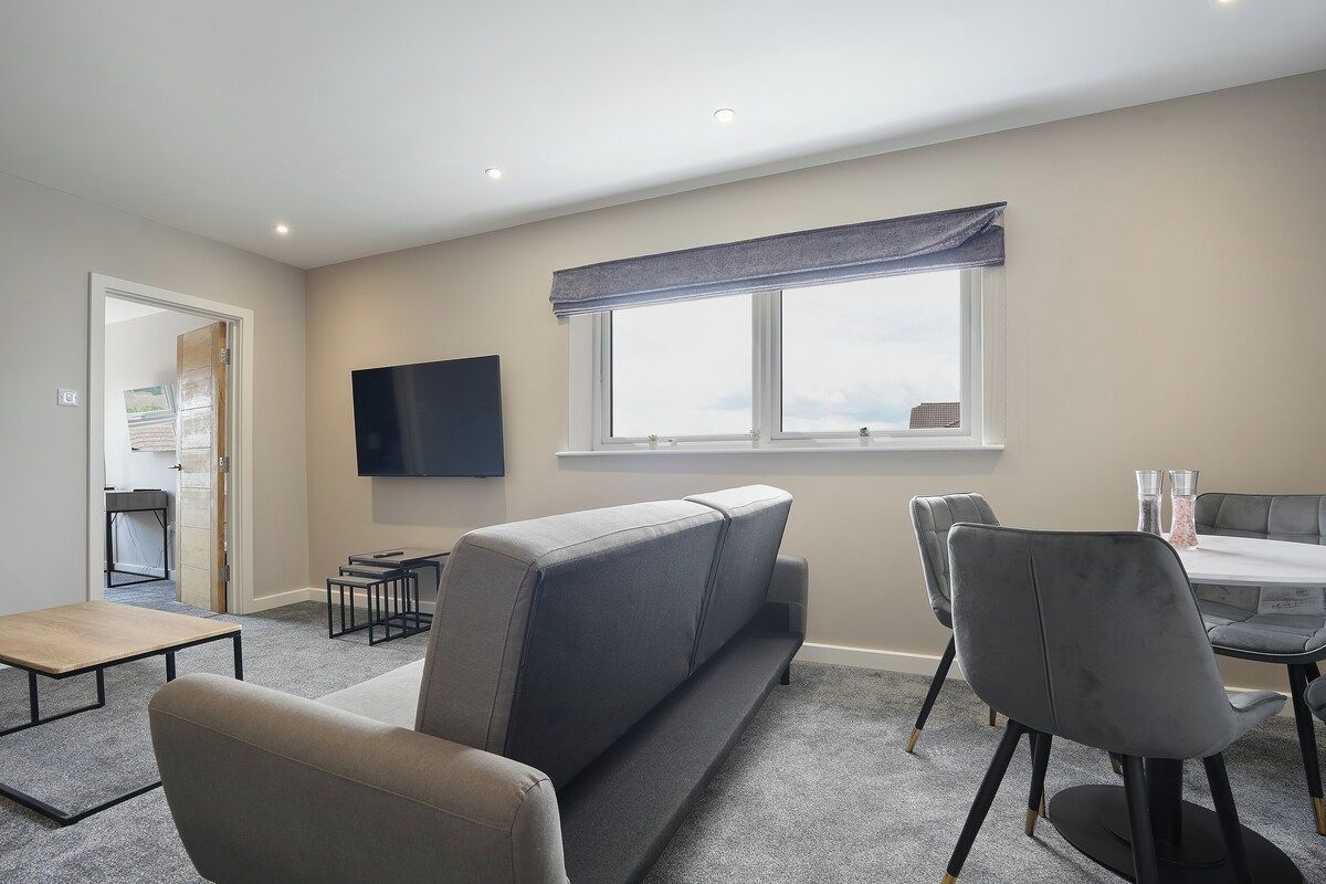 A modern living room with a sofa, a dining table, and chairs. There is a flat-screen TV mounted on the wall and a window with a view.