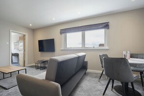 A modern living room with a sofa, a dining table, and chairs. There is a flat-screen TV mounted on the wall and a window with a view. - The Olive Tree | Deluxe 1-Bed Apartment (Kingston upon Hull)
